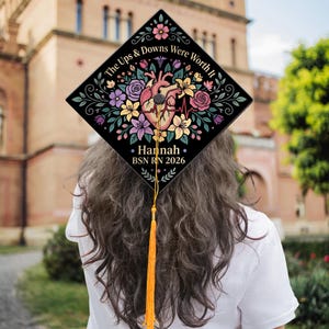 May include: A black graduation cap with floral and anatomical heart designs. The cap reads "The Ups & Downs Were Worth It" and includes the name "Hannah" and "BSN RN 2026". A gold tassel hangs from the cap.