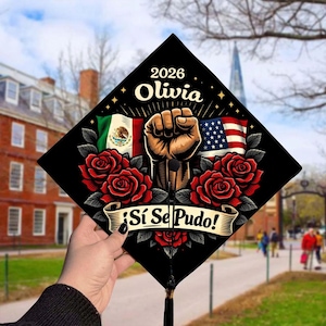 May include: Black graduation cap decorated with a raised fist, red roses, and the flags of Mexico and the United States. The cap includes the name "Olivia" and the year "2026". A banner reads "¡Sí Se Pudo!"