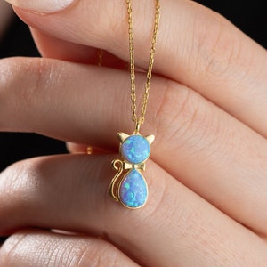 May include: A gold chain necklace with a blue opal cat pendant. The cat is sitting with its tail curled around its body.