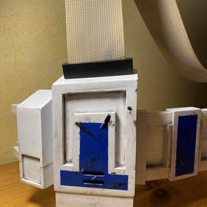 Peut inclure: Une ceinture en plastique blanche et bleue avec une boucle noire et une sangle beige. La ceinture a un aspect usé et vieilli, avec des éclaboussures de peinture noire et des rayures.