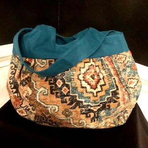 May include: A large tote bag with a teal suede top and strap. The lower portion of the bag features a patterned fabric in shades of brown, orange, and teal, with an intricate geometric design. The bag is sitting on a black surface.