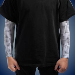 May include: A black short-sleeved t-shirt worn over a pair of arm sleeves featuring a white, grey, and black geometric grid pattern. The sleeves extend from the wrists to the upper arms, contrasting with the solid black fabric of the t-shirt.