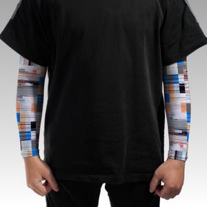 May include: A black short-sleeved t-shirt worn over long sleeves featuring a white base with a digital glitch pattern in shades of blue, orange, and black. The garment has a solid black torso and a multi-colored geometric print on the sleeves.