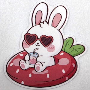 Op de afbeelding: Een gestanste sticker met een wit cartoonkonijn met roze wangen en een rode hartvormige zonnebril. Het konijn zit in een rode opblaasbare zwemband met aardbeienpatroon en houdt een klein glas met een roze drankje en een rietje vast.