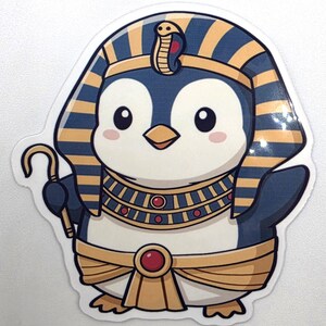 May include: A die-cut sticker featuring a cartoon penguin dressed as an Egyptian pharaoh. The penguin wears a blue and gold striped nemes headpiece with a cobra, a wide beaded collar with red accents, and a gold waist wrap with a red gem.