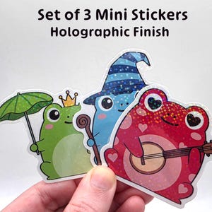 May include: Set of 3 mini stickers with a holographic finish. The stickers feature cartoon frogs: one green with an umbrella, one blue with a wizard hat, and one red playing a banjo. Text on the image reads "Set of 3 Mini Stickers Holographic Finish."