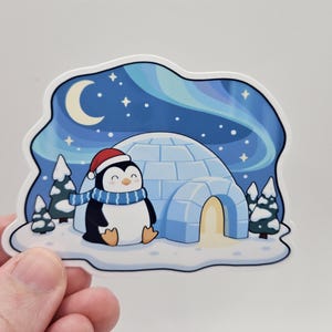 May include: A sticker featuring a cartoon penguin wearing a red hat and blue scarf, sitting in front of an igloo. The background shows a night sky with the aurora borealis, a crescent moon, and stars. Evergreen trees are also present.
