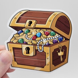 May include: A sticker of a cartoon treasure chest, overflowing with gold coins, colorful gems, and a pearl necklace. The chest is brown with gold trim and a keyhole. A hand is holding the sticker.