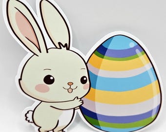 May include: A die-cut sticker featuring a light cream-colored cartoon rabbit with long ears and pink cheeks, standing next to a large, glossy egg decorated with horizontal stripes in shades of blue, yellow, green, and white against a plain background.