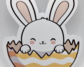 May include: A die-cut sticker featuring a white bunny with pink cheeks emerging from a cracked, yellow and white wavy-patterned egg. The bunny has long ears with pink inner details, a small black nose, and closed eyes, set against a plain background.