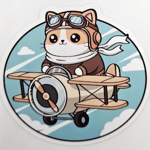 May include: A sticker featuring a cartoon cat wearing a pilot's helmet and scarf, flying a biplane. The cat has brown and white fur and large eyes. The biplane is beige and brown, set against a blue sky background.