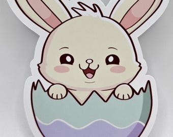 May include: A die-cut sticker featuring a cartoon rabbit with cream-colored fur, large dark eyes, and pink cheeks, emerging from a cracked eggshell. The eggshell is colored in light teal and lavender with a wavy line pattern separating the two colors.