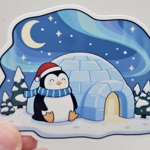 May include: A die-cut sticker featuring a cartoon penguin wearing a red Santa hat and a blue striped scarf. The penguin sits in front of a blue ice igloo with a glowing entrance, surrounded by snow-covered pine trees under a night sky with a crescent moon, stars, and blue aurora borealis.