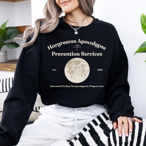 May include: Black sweatshirt with a white graphic design that says "Hargreeves Apocalypse Prevention Services" with a moon and an umbrella graphic. The text also says "Est. 2019" and "Guaranteed To Stop The Apocalypse In 7 Days or Less!"