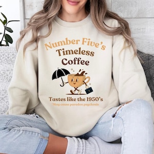 May include: A beige sweatshirt with the text "Number Five's Timeless Coffee" and a cartoon coffee cup holding an umbrella and a briefcase. The text below the image reads "Tastes like the 1950's *May cause paradox psychosis*"