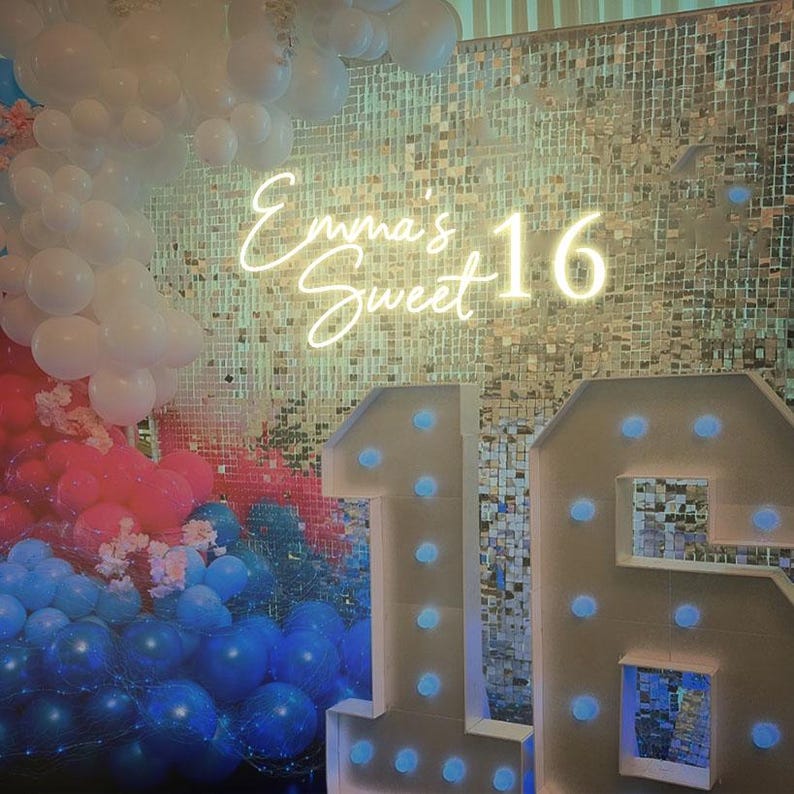 May include: A white backdrop with a silver sequin background. The words "Emma's Sweet 16" are in white neon lights. There are two large white numbers, 1 and 6, with white lights around the edges. The numbers are decorated with blue balloons.