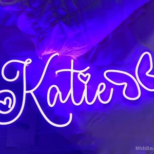 May include: A neon sign in white script spelling "Katie" with a heart detail. The sign is illuminated with a bright white light against a dark purple background. The sign is a decorative item.