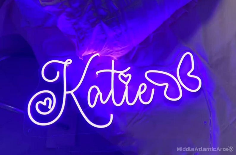 May include: A neon sign in white script spelling "Katie" with a heart detail. The sign is illuminated with a bright white light against a dark purple background. The sign is a decorative item.