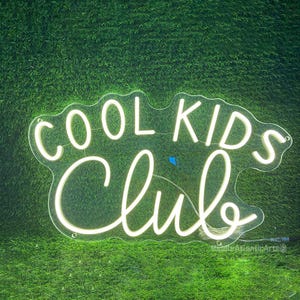 May include: White neon sign with the words "Cool Kids Club" in a cursive font. The sign is on a green grassy background.