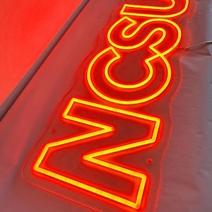 Puede incluir: Un letrero de neón con la palabra "NCU" en letras de neón amarillas sobre un fondo de acrílico transparente. El letrero está encendido y brilla en rojo.