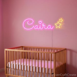 May include: A pink nursery with a wooden crib and a neon sign that spells out the name "Caira" in cursive. The sign includes a heart and two yellow stars. The wall is painted a soft pink.