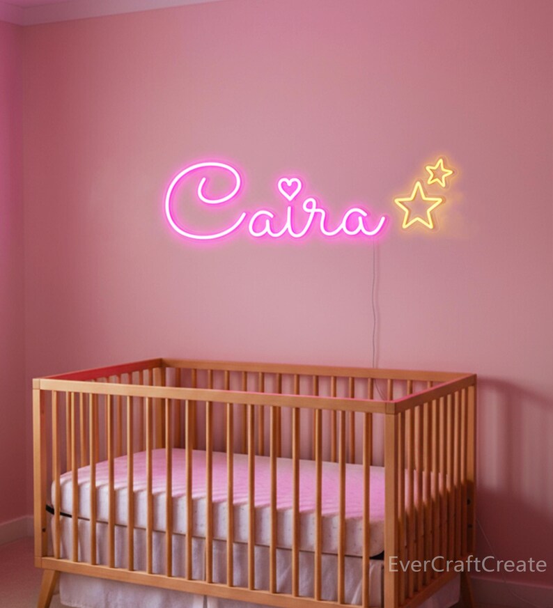May include: A pink nursery with a wooden crib and a neon sign that spells out the name "Caira" in cursive. The sign includes a heart and two yellow stars. The wall is painted a soft pink.