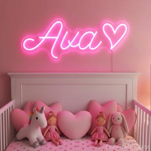 May include: A pink neon sign spelling "Ava" with a heart symbol, mounted above a crib. The crib is decorated with heart-shaped pillows, dolls in pink dresses, a unicorn, and a bunny. The bedding has a pink heart pattern.