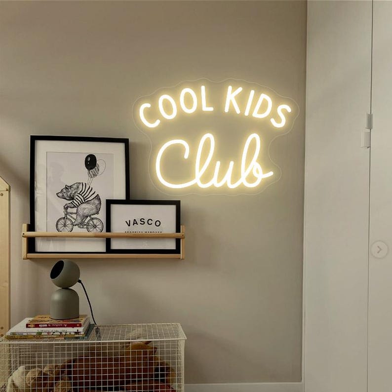 May include: White neon sign with the text "COOL KIDS CLUB" on a beige wall. The sign is hanging above a wooden shelf with two framed prints.