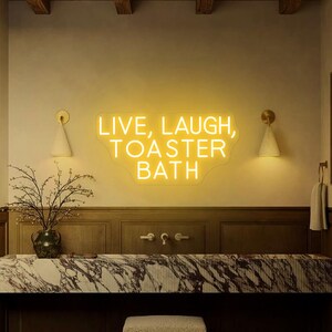 Puede incluir: Un letrero de ne&oacute;n amarillo con el texto "Live, Laugh, Toaster Bath" en una pared blanca en un ambiente de ba&ntilde;o.