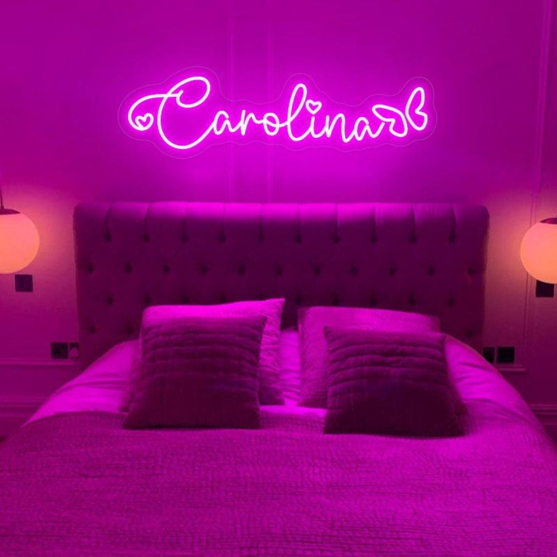 May include: A neon sign in a bedroom spells out the name "Carolina" in a cursive font, with a heart symbol. The sign is mounted above a bed with a tufted headboard and two decorative pillows. The room is illuminated with a pink glow.