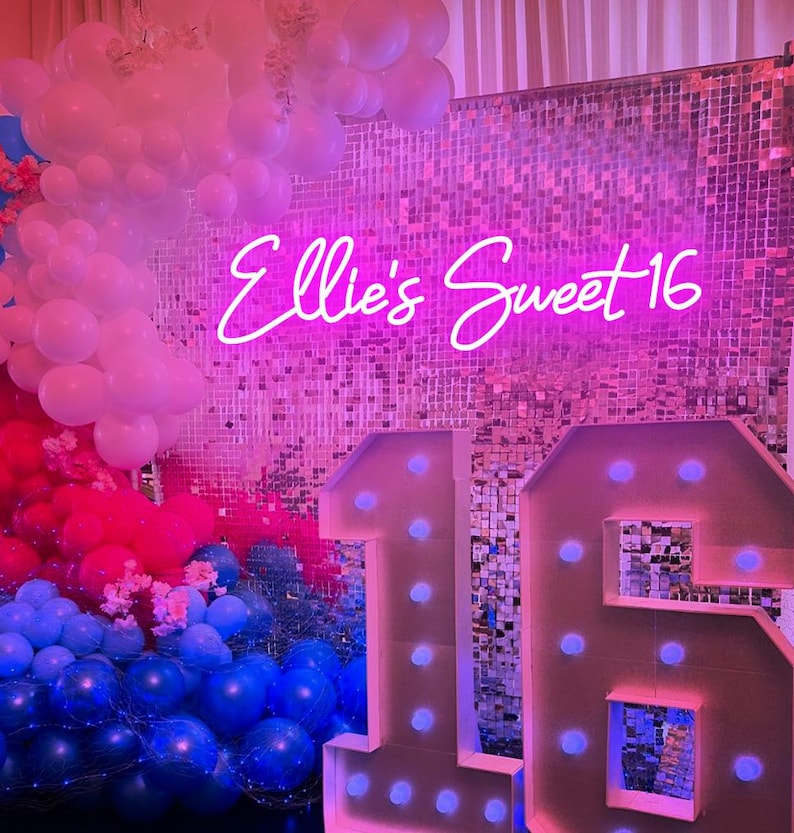 May include: A pink and silver sequin backdrop with a neon sign that says "Ellie's Sweet 16" and large white light-up numbers 1 and 6. The backdrop is decorated with pink, blue, and white balloons.
