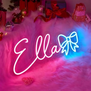 May include: A neon sign in a cursive font spelling "Ella" with a bow design. The sign glows with a gradient of pink and blue light. The sign is placed on a fluffy, pink surface, with Christmas decorations in the background.