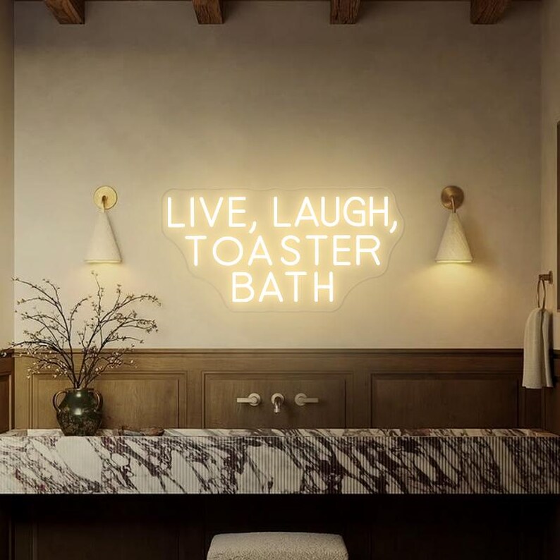 Puede incluir: Un letrero de ne&oacute;n blanco con el texto "LIVE, LAUGH, TOASTER BATH" en una pared blanca en un ambiente de ba&ntilde;o. El letrero est&aacute; iluminado y tiene un borde ondulado.