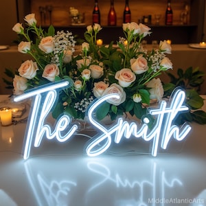 Puede incluir: Un letrero de neón que dice "The Smith" en una fuente cursiva, iluminado con una luz blanca brillante. El letrero se coloca frente a un arreglo floral de rosas rosas y vegetación, creando un ambiente romántico.