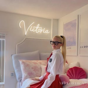 May include: A bedroom scene featuring a white neon sign that reads "Victoria". The room has a bed with a tufted headboard, pillows, and a red skirt. The person is wearing a white shirt, a red tie, and black glasses.