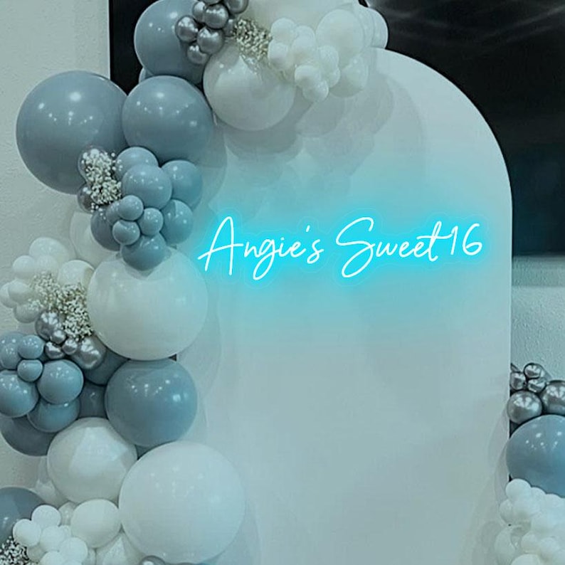 May include: A white backdrop with a blue neon sign that reads "Angie's Sweet 16". The backdrop is decorated with a garland of white, gray, and silver balloons.