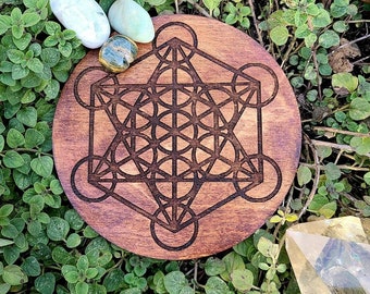 May include: A round wooden disc with a dark brown stain and a laser-etched geometric pattern, known as the Flower of Life, in black. Three small stones are placed on the disc.