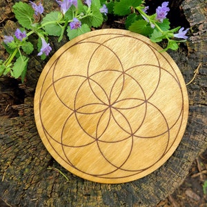 Peut inclure: Un objet d&eacute;coratif rond en bois clair, orn&eacute; d'un motif de Fleur de Vie grav&eacute; au laser en brun fonc&eacute;. Le bois clair contraste avec les lignes grav&eacute;es. Pos&eacute; sur une souche d'arbre vieillie, avec des fleurs violettes et des feuilles vertes.