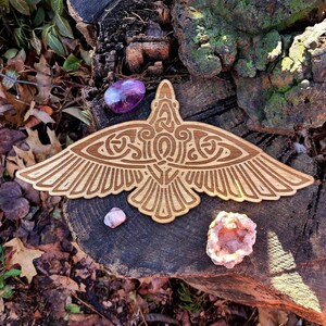 May include: A wooden carving of a stylized raven with intricate details. The raven is spread-winged and has a Celtic knot design on its body. The carving is on a natural wood background with small pink and purple crystals.