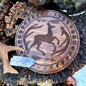 May include: A wooden round coaster with an engraved design of a deer with antlers, surrounded by a circle of moon phases. The coaster is brown and has a natural wood finish.
