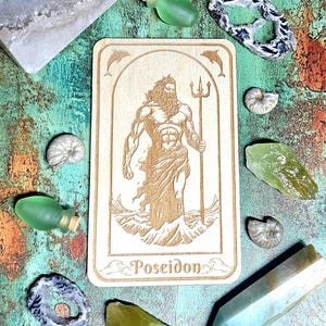 Könnte beinhalten: Eine Holzkarte mit dem Wort "Poseidon" und einem gravierten Bild des griechischen Gottes Poseidon mit einem Dreizack. Das Bild ist von Kristallen, kleinen grünen Flaschen und Muscheln umgeben. Der Hintergrund ist türkis und kupferfarben.