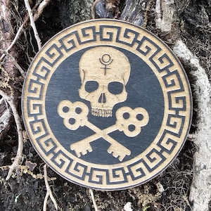 May include: A circular wooden plaque with a skull and crossed keys design. The skull is in the centre on a black background, framed by a Greek key pattern. The piece is made from light and dark wood.
