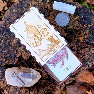 Könnte beinhalten: Eine Holzkiste mit einem Scharnierdeckel, die ein Tarotkartenspiel enthält. Die Kiste ist mit einem Design einer nach oben zeigenden Hand verziert, mit den Worten "The Finger" und "Stones" aufgedruckt. Die Kiste ist geöffnet und zeigt die Karten im Inneren.