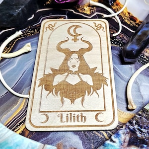 May include: A wooden tarot card featuring an engraved illustration of Lilith with horns, a crescent moon, and the text "Lilith C". The card is rectangular with rounded corners. The artwork is in a light brown color.