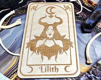 May include: A wooden tarot card featuring an engraved illustration of Lilith with horns, a crescent moon, and the text "Lilith C". The card is rectangular with rounded corners. The artwork is in a light brown color.
