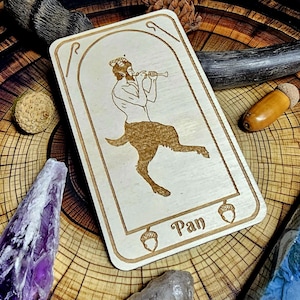 May include: A wooden card featuring an engraved illustration of Pan playing a flute, with a goat's lower body. The card is surrounded by crystals, acorns, and other natural elements, set on a wood-grain surface.