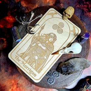K&ouml;nnte beinhalten: Eine Tarotkarte aus Holz mit dem Text "The Morrigan" und einer eingravierten Illustration einer Frau, die einen Speer h&auml;lt. Die Karte ist von Kristallen, einem Sch&auml;del, einem Vogelsch&auml;del und einem dekorativen Metallst&uuml;ck auf einem Holzsockel umgeben.