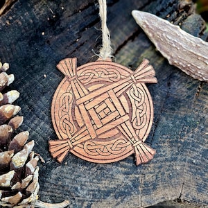 Könnte beinhalten: Runder Holzschmuck mit keltischem Knotenmuster. Der hellbraune Schmuck hat eine Schnurschlaufe zum Aufhängen. Das Design zeigt verschlungene Muster und ein zentrales Quadrat. Er wird auf einer dunklen, strukturierten Oberfläche präsentiert.
