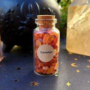 Puede incluir: Una pequeña botella de vidrio llena de cristales de cornalina naranja. La botella tiene un tapón de corcho y una etiqueta de papel marrón que dice "Cornalina".