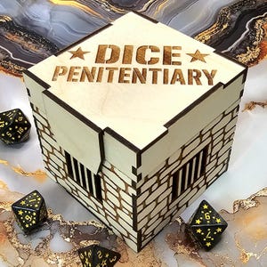May include: A square wooden box designed like a prison cell with stone wall patterns and barred windows. The lid is engraved with the text DICE PENITENTIARY and two stars. Three black polyhedral dice with gold star numbering are placed nearby.